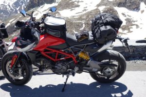 Ein rot-schwarzes Motorrad mit Reisetaschen steht auf einer Bergstraße, Schneeflecken übersäen die felsige Landschaft.