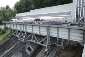 Ein großes Boot fährt durch ein Kanallift, während Zuschauer zwischen Bäumen und einer modernen Gebäudekulisse zuschauen.
