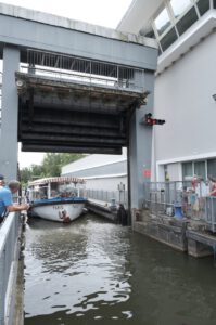 Ein Boot namens Paris erregt Aufmerksamkeit, während es zwischen modernen Stadtgebäuden und neugierigen Zuschauern durch eine Kanalschleuse navigiert.
