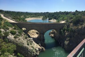 Eine dreibogige Steinbrücke überquert anmutig einen Fluss inmitten von Klippen und Grün und führt zu einem ruhigen See.