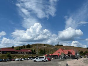 Vor Gebäuden mit roten Dächern befindet sich ein Parkplatz mit Autos und Motorrädern, im Hintergrund grasbewachsene Hügel und Wolken.