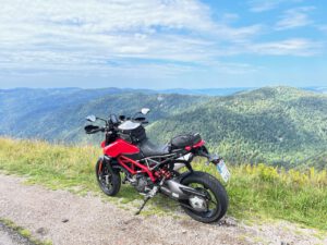 Ein rotes Motorrad steht auf einem Schotterweg und bietet einen atemberaubenden Blick auf grüne Hügel unter einem teilweise bewölkten Himmel.