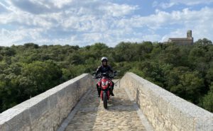 Ein behelmter Fahrer fährt mit einem roten Motorrad über eine Steinbrücke, umgeben von üppigem Grün und einer Kirche in der Ferne.