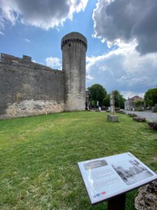 Neben einer Mauer steht ein historischer Steinturm mit Zinnen. In der Nähe befindet sich ein grüner Rasen und eine Informationstafel.