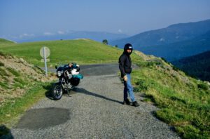 Ein Fahrer mit Helm und dunkler Jacke steht neben seinem Motorrad auf einer Straße, die sich perfekt für geführte Touren eignet, vor ihm liegen Berge.