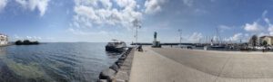 Ein Steinpier mit angedockten Booten, ein grüner Leuchtturm und Gebäude unter einem ruhigen, teilweise bewölkten Himmel.