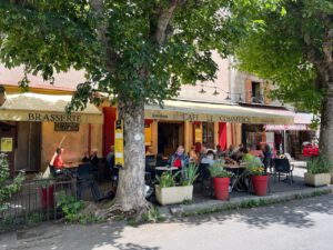 Eine charmante Café-Szene mit Sitzgelegenheiten im Freien unter Bäumen, bewundert von vorbeifahrenden Motorradreisegruppen.