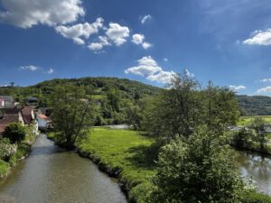 Ein ruhiger Fluss fließt an einem grünen Feld vorbei, das mit Bäumen und Hügeln dahinter perfekt für geführte Motorradtouren ist.