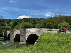 Zwei Radfahrer halten auf einer Steinbrücke mit drei Bögen inne, umgeben von üppigen Hügeln und lebendiger Natur.