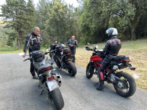 Drei Motorradfahrer in schwarzer Montur halten auf einer von Bäumen gesäumten Straße an und unterhalten sich neben ihren abgestellten Motorrädern.