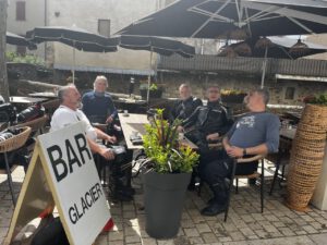 Fünf Männer unterhalten sich in einem Straßencafé über geführte Motorradreisen. Das Schild der Bar Glacier und Steingebäude schmücken die Szene.