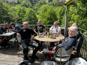 Eine Gruppe in Motorradmontur entspannt sich bei Drinks auf einem Deck im Grünen und lächelt neben ihren Helmen.