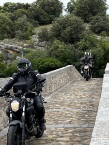 Zwei Motorradfahrer in Schwarz fahren dicht an dicht über eine Steinbrücke, umgeben von üppigem Grün, auf einer malerischen Tour.