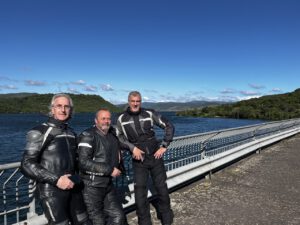 Drei Biker genießen eine geführte Tour und genießen die Aussicht auf den See und die grünen Hügel unter einem klaren blauen Himmel.