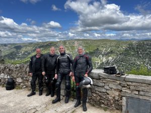 Vier Radfahrer in voller Montur genießen lächelnd am Steingeländer einen malerischen Aussichtspunkt mit Hügeln und Wolken.