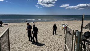 **Menschen schlendern an einem sonnigen Strand mit ruhiger See entlang. Links ein offenes Tor, rechts versammeln sich Biker.**