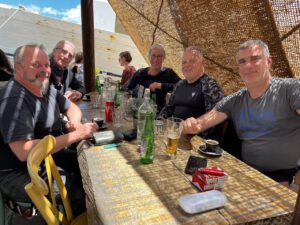 Fünf Personen genießen Getränke an einem Holztisch, während das Sonnenlicht durch das geflochtene Dach über ihnen tanzt.