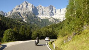 Ein Motorradfahrer unternimmt eine geführte Bergtour, flankiert von einem Auto, üppigen Bäumen und felsigen Gipfeln.