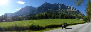 Ein Motorrad steht auf einer Wiese, dahinter majestätische Berge, unter einem klaren Himmel, auf den das Sonnenlicht strömt.