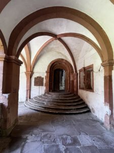 Ein mittelalterlicher Steinflur mit Bögen imitiert eine Motorradtour, Stufen führen zu einer geschnitzten Holztür.