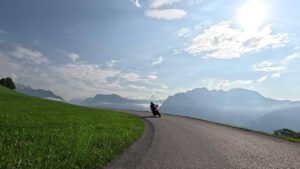 Ein Motorradfahrer lehnt sich in eine Kurve, flankiert von üppigem Gras und dunstigen Bergen unter einem wolkenverhangenen Himmel.