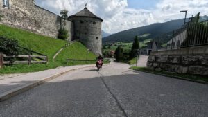 Ein Motorradfahrer fährt auf einer Panoramastraße an einem Steinturm vorbei, umgeben von grünem Gras und Bergen in der Ferne.