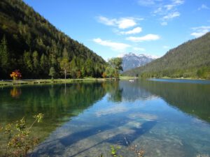 In einem ruhigen See spiegeln sich Himmel und Kiefern, in der Ferne ist ein schneebedeckter Gipfel zu sehen – ein malerischer Traum für jeden Biker.