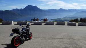 Ein Motorrad an einem Aussichtspunkt mit drei Personen, die unter klarem Himmel die Aussicht auf Berge und See genießen.