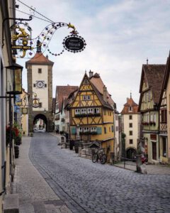Eine Kopfsteinpflasterstraße mit bunten Fachwerkhäusern führt unter bedecktem Himmel zu einem historischen Steinturm.