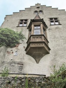 Ein reich verzierter Erker ziert die Steinfassade mit Efeu und Spitzbögen unter einem bewölkten Himmel.