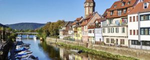 Ein von bunten Häusern und Booten gesäumter Kanal mit einem historischen Turm, üppigen Bäumen und grünen Hügeln in der Ferne.