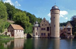 Ein Schloss mit rundem Turm spiegelt sich in einem See, flankiert von grünen Bäumen und einem Steingebäude mit rotem Dach.