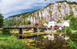 Ein Dorf mit rot-weißen Häusern, einer Holzbrücke über einem ruhigen Fluss, umrahmt von felsigen Klippen und üppigem Wald.