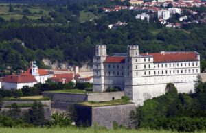 Ein historisches Schloss mit Zwillingstürmen im Grünen, vor der Kulisse einer Stadt, eines Waldes und ferner Hügel.