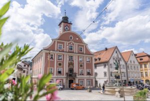 Historischer Uhrturm mit pastellfarbener Fassade, kunstvollen Details, Brunnen und blühenden Pflanzen unter einem bewölkten Himmel.