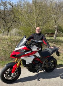 Eine Person in einer schwarz-roten Jacke steht neben einer roten Ducati auf einer Straße, umgeben von Bäumen unter klarem Himmel.