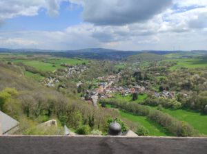 Ein kleines Dorf liegt in einem üppig grünen Tal mit sanften Hügeln, Wäldern und einem teilweise bewölkten Himmel über sich.