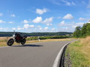 Ein schwarzes Motorrad steht neben einer kurvenreichen Landstraße inmitten von üppigen Feldern, Hügeln und einem strahlend blauen Himmel.