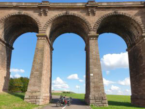 Ein steinernes Viadukt mit drei Bögen überspannt eine Landstraße; darunter ist ein rot-schwarzes Motorrad geparkt.