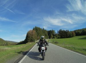 Ein Motorradfahrer in schwarzer Kleidung und mit Helm fährt eine leere Landstraße entlang, flankiert von Feldern und Bäumen.