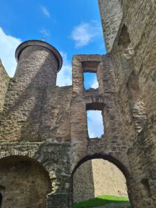Alte Burgruine aus Stein mit Bögen, hohen Mauern und einem runden Turm unter blauem Himmel, darunter Gras.