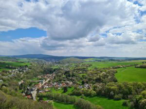 Ein malerisches Dorf, eingebettet zwischen üppigen Feldern, sanften Hügeln und Wäldern unter einem hellen, wolkengesprenkelten Himmel.