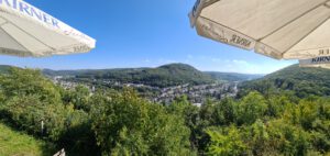 Panoramablick von einer Terrasse mit weißen Kirner-Sonnenschirmen auf ein grünes Tal und die darunter liegende Stadt.