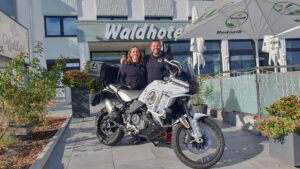 Zwei Personen lächeln hinter einem weißen Abenteuer-Motorrad vor dem Waldhotel, mit Pflanzen und einem großen Regenschirm.