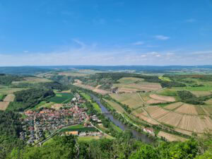 Ein gewundener Fluss fließt vorbei an Feldern, Wäldern und einem kleinen Dorf unter einem blauen, wolkenverhangenen Himmel.