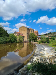Ein steinernes Gebäude am Ufer eines Flusses, das sich im ruhigen Wasser spiegelt, mit Bäumen und einem Wasserfall unter blauem Himmel.