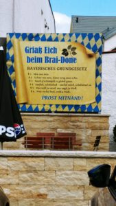 Ein deutsches Schild mit bayerisch-blau-weißem Muster listet lustige Sprüche als Grundgesetz auf; davor ein Pepsi-Schirm.