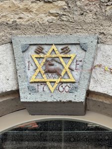 Eine Steintafel über einer Tür zeigt einen gelben Davidstern, ein Lamm in der Mitte, Hände, "1760" und "I E".