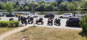 Motorräder und Autos sind entlang einer Uferstraße geparkt, Menschen in der Nähe, Bäume und Häuser dahinter.