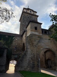 Historischer Steinturm mit Efeu und Bögen, der durch den Haupttorbogen auf den Kopfsteinpflasterweg hinaufleuchtet.